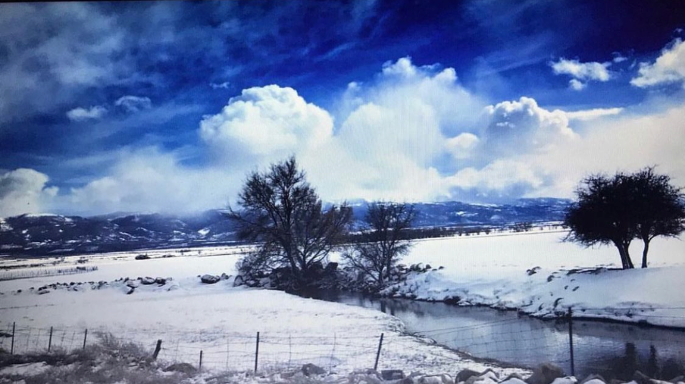 A brook curves through snow beneath a blue sky, with bare trees, a foreground fence, and distant hills.
