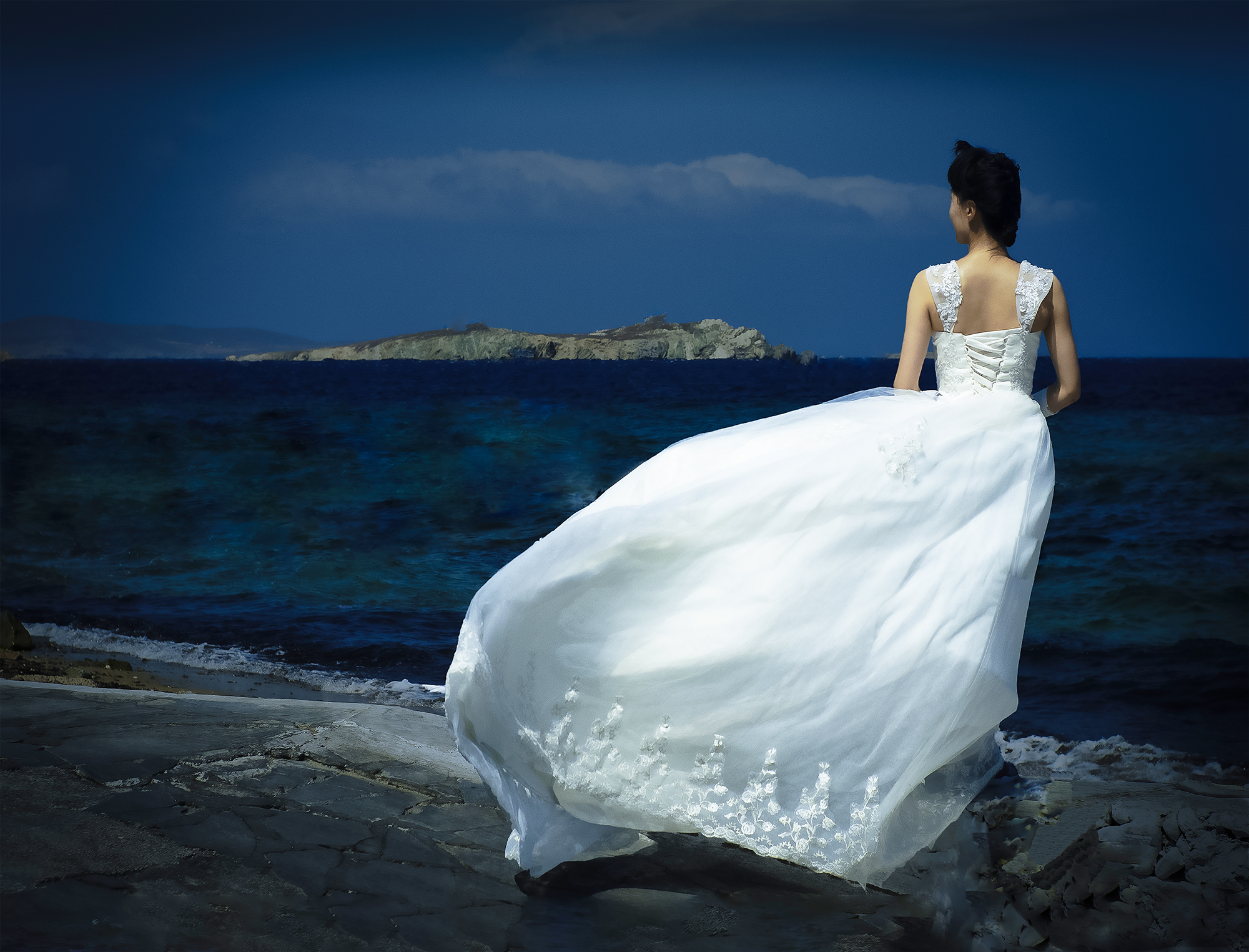 Gene Sellers: Photographs at the Edge of a Story | ArtTour International A woman in a flowing white dress faces a dark blue sea and distant island, with the dress extending across a rocky foreground.